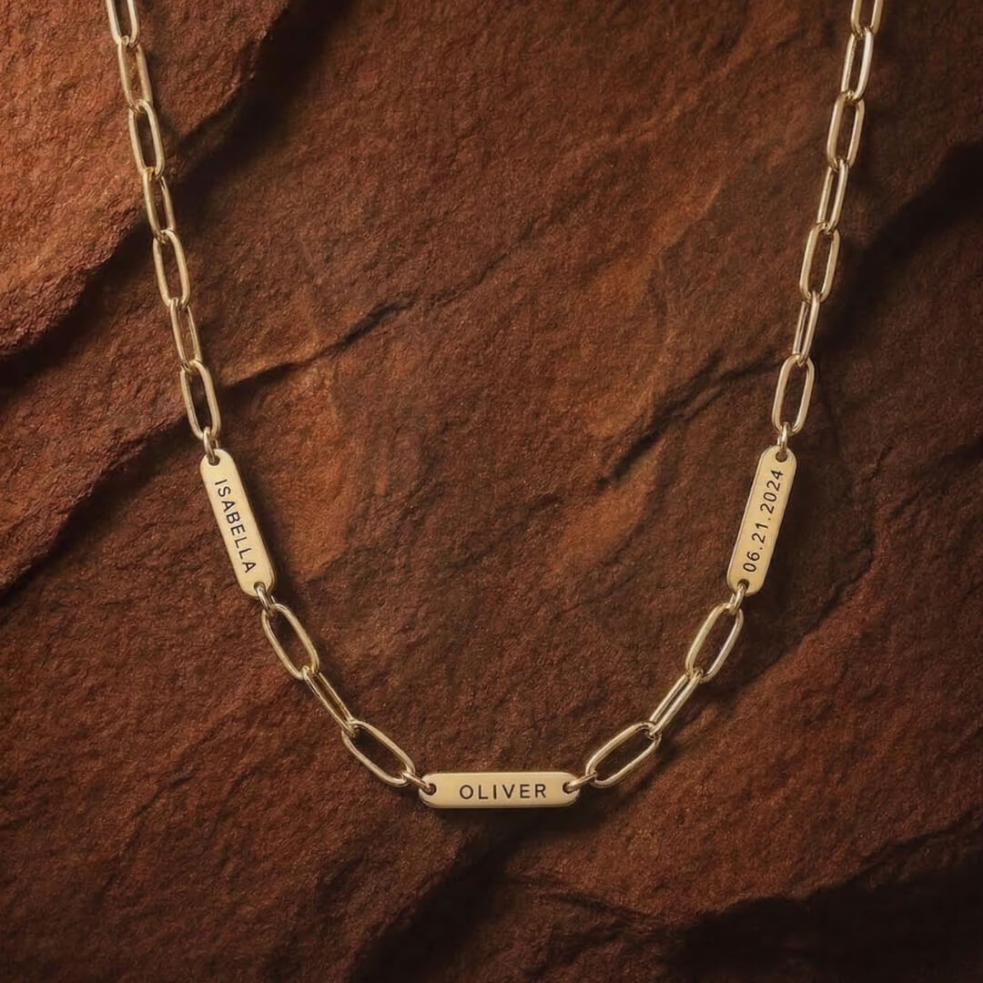 JUNE ELLE gold engraved tag name necklace with Isabella, Oliver and 06.21.2024 on paperclip chain front view on chocolate textured background