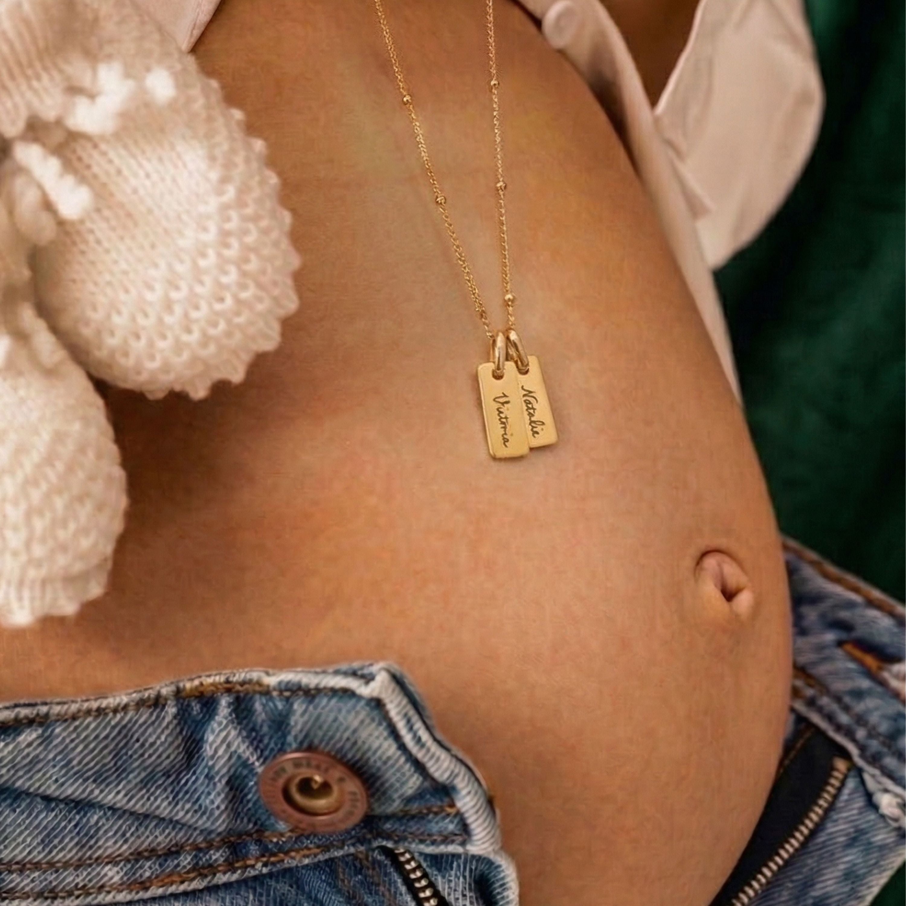 JUNE ELLE Gold Engraved Bar Necklace featuring two personalized nameplates, 'Victoria' and 'Natalie', layered on a satellite chain, held by hand with pendants dangling over the baby bump.