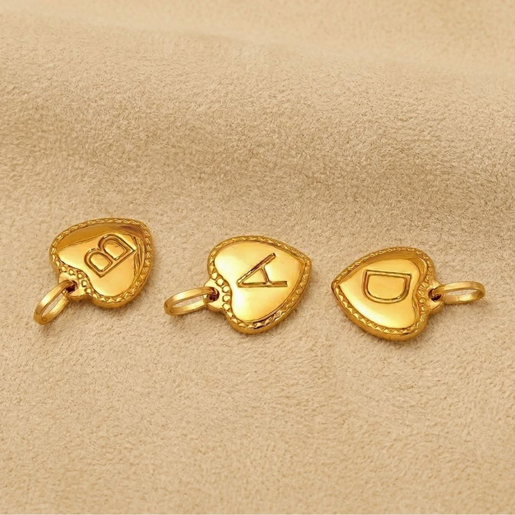 JUNE ELLE gold heart initial charm letters A B and D laid out on light brown fabric background
