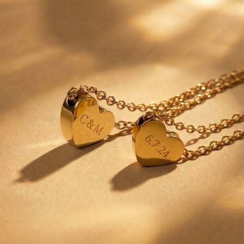 JUNE ELLE two gold custom heart necklaces engraved with 6.7.24 and C&M displayed upright on light brown fabric with soft lighting