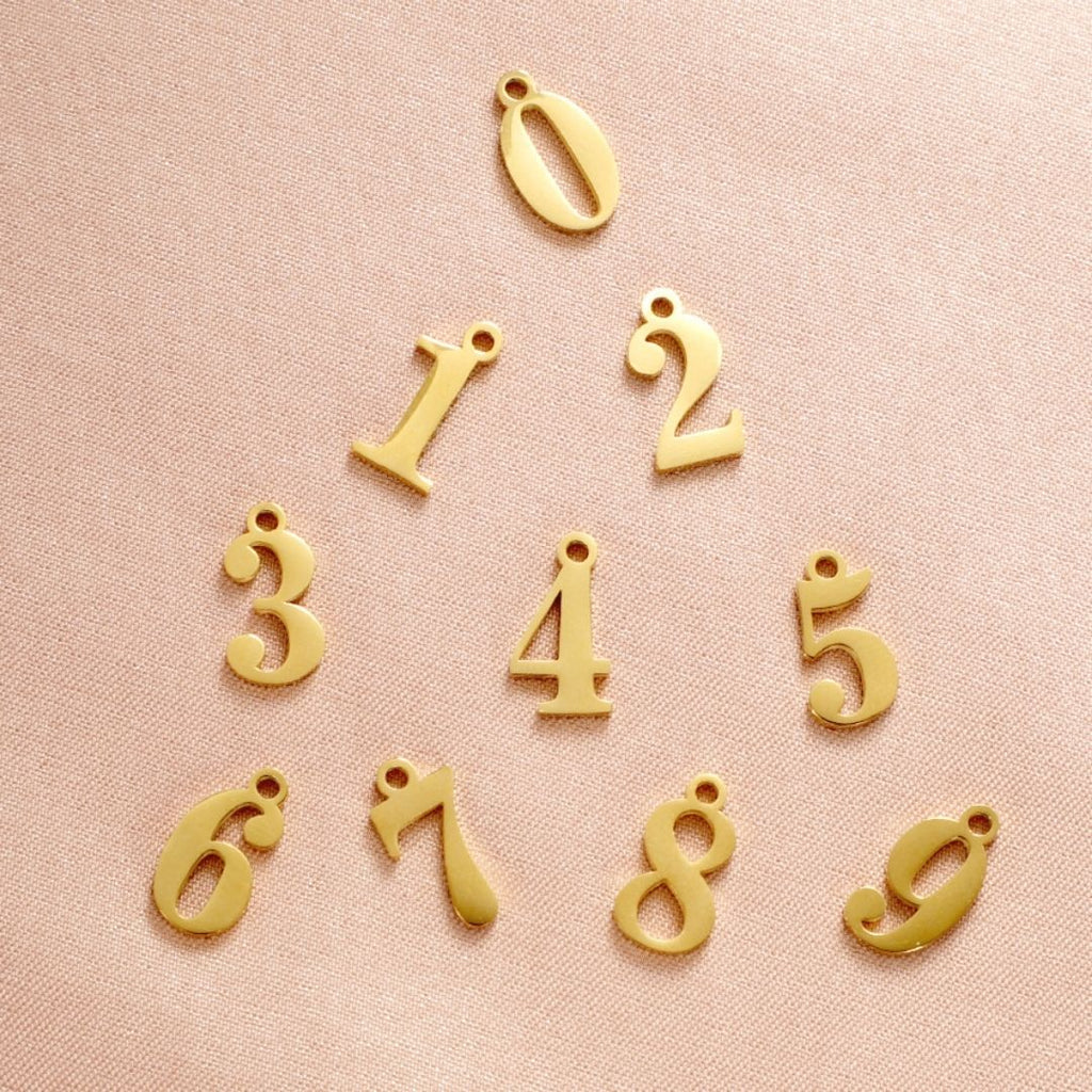 JUNE ELLE gold lucky number charm numbers 0–9 arranged on pink background
