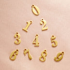 JUNE ELLE gold lucky number charm numbers 0–9 arranged on pink background