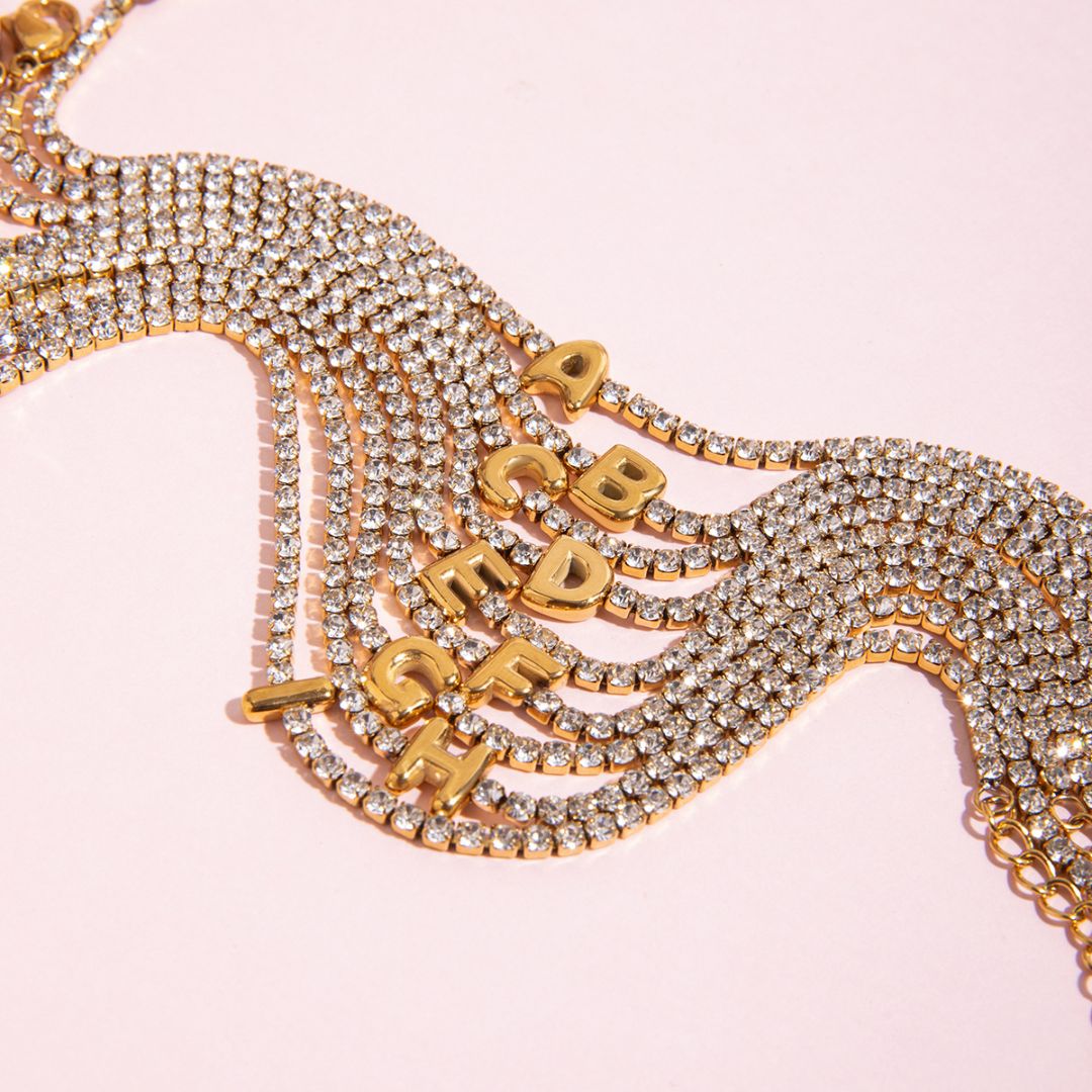 JUNE ELLE gold initial tennis bracelet featuring letters A to I arranged in a casual clustered layout on pink background
