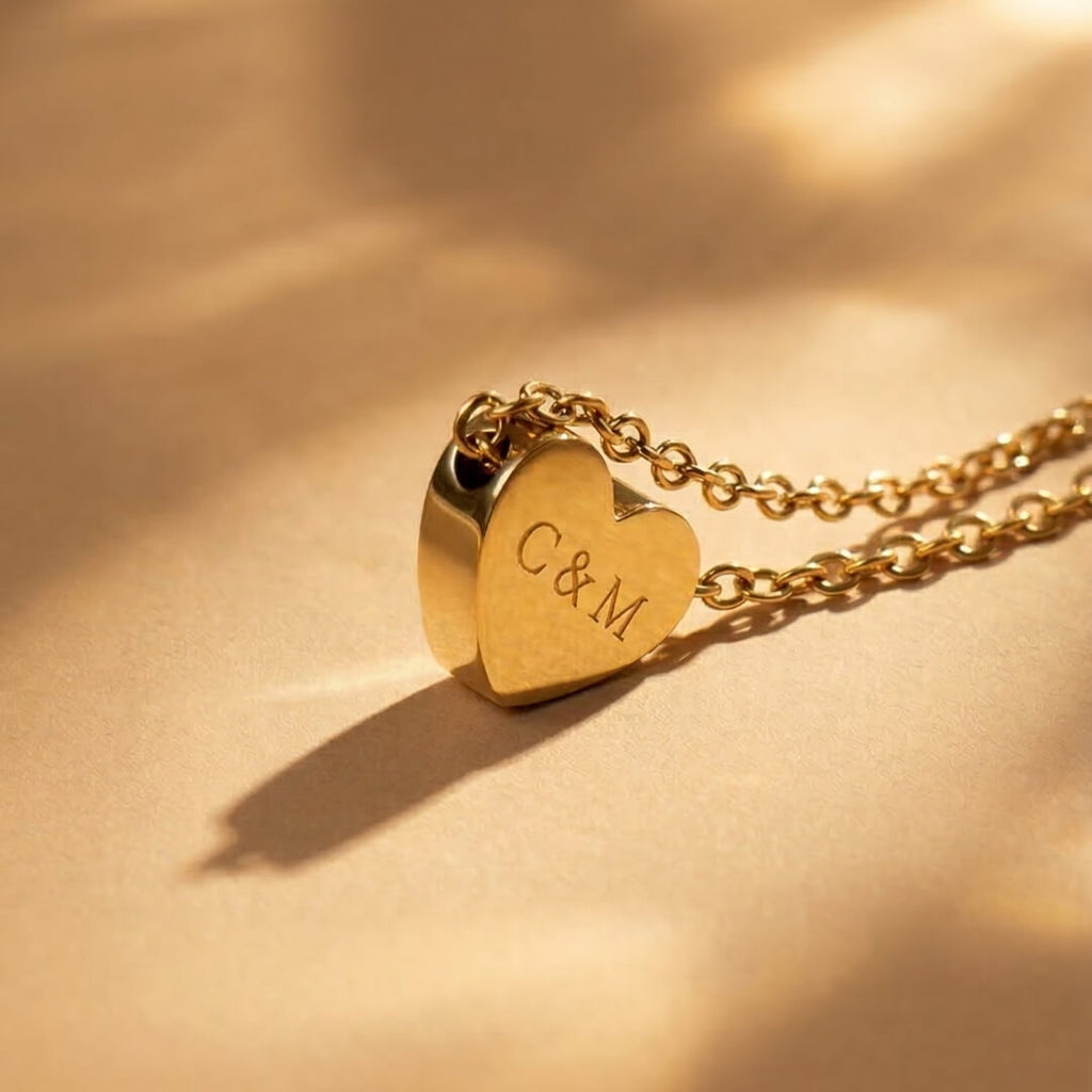 JUNE ELLE gold custom heart necklace engraved with C&M displayed upright on light brown fabric with soft lighting