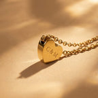 JUNE ELLE gold custom heart necklace engraved with C&M displayed upright on light brown fabric with soft lighting