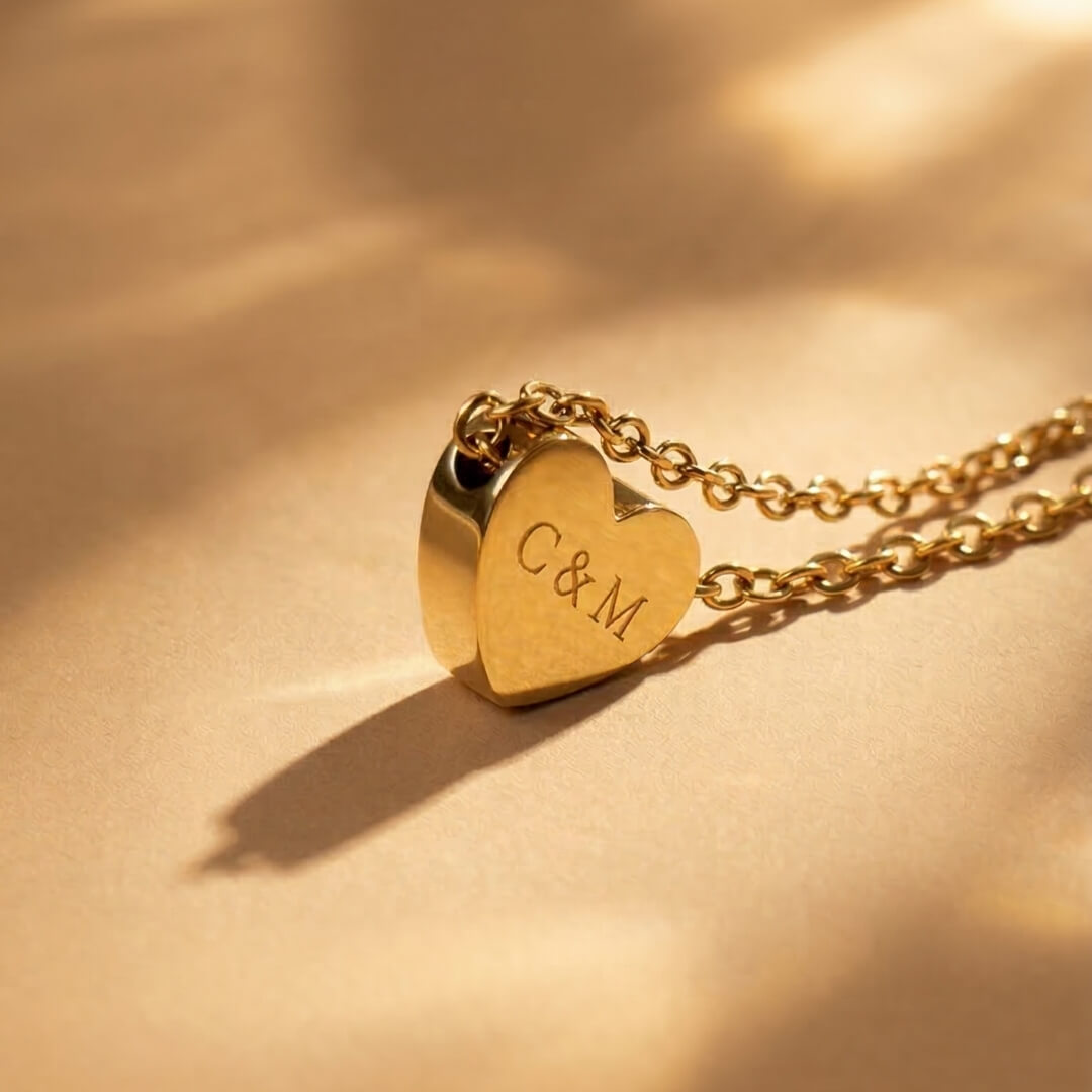 JUNE ELLE gold custom heart necklace engraved with C&M displayed upright on light brown fabric with soft lighting