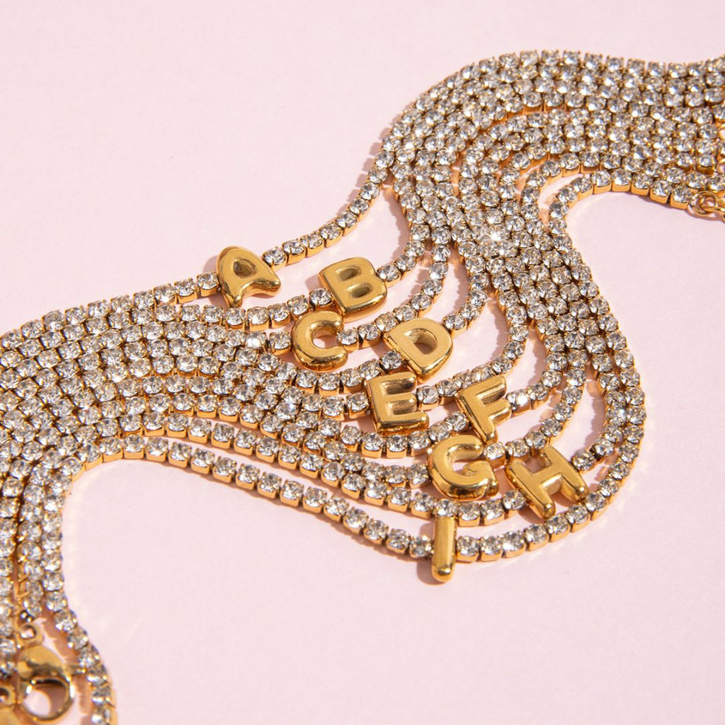 JUNE ELLE gold initial tennis bracelet featuring letters A to I arranged in a casual clustered layout on pink background with soft composition framing