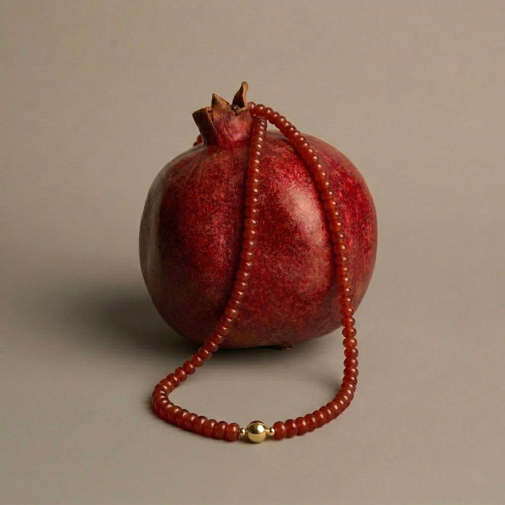 JUNE ELLE MagMix Carnelian Bead Necklace with a central gold magnetic clasp, gracefully draped over a ripe, dark red pomegranate on a neutral background.