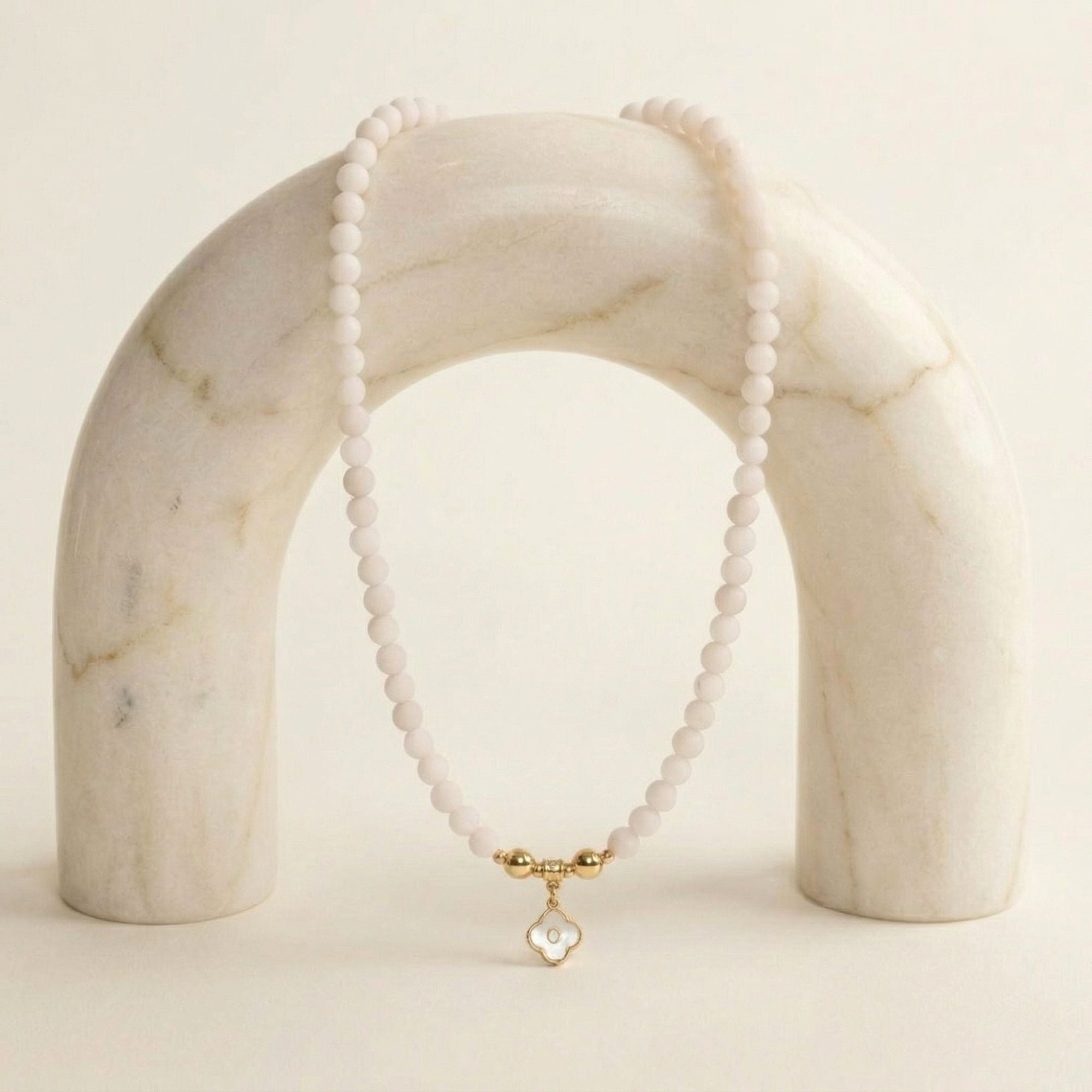JUNE ELLE MagMix Rose Quartz Bead Necklace connected to a custom gold mother-of-pearl clover initial charm via magnetic clasps, gracefully draped over a marble arch prop.