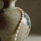 Close-up detail of the JUNE ELLE MagMix seed freshwater pearl necklace, showcasing lustrous oval pearls separated by delicate gold spacer beads against a textured ceramic background.