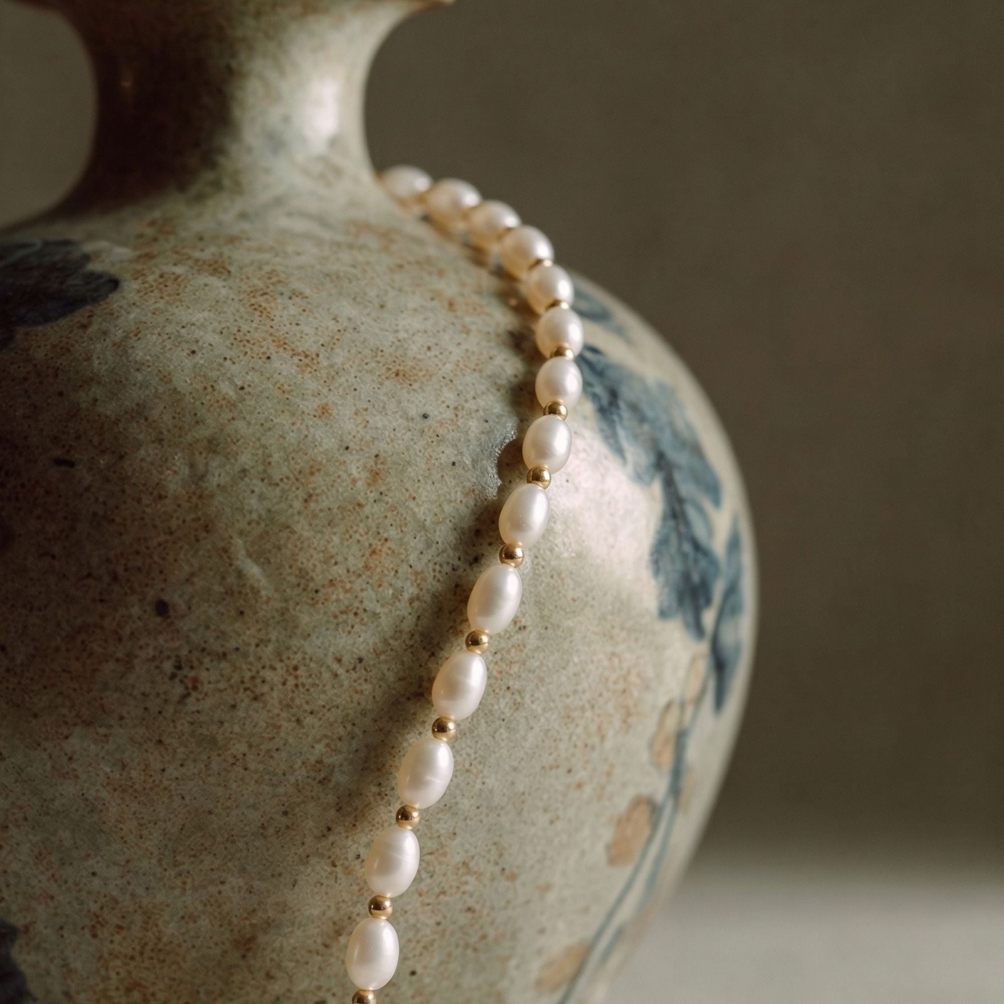 Close-up detail of the JUNE ELLE MagMix seed freshwater pearl necklace, showcasing lustrous oval pearls separated by delicate gold spacer beads against a textured ceramic background.