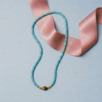JUNE ELLE MagMix Turquoise Bead Necklace connected to a MagMix Tiger’s Eye Barrel Centerpiece via a magnetic clasp, styled with a pink ribbon on a light blue background.