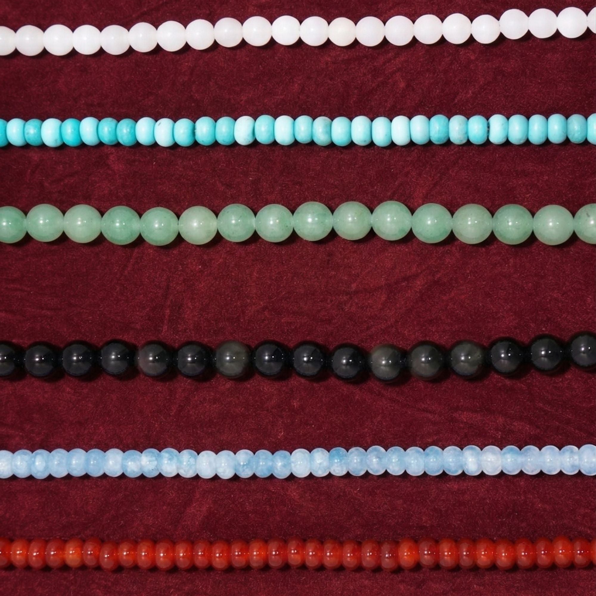 Flat lay display of the JUNE ELLE MagMix interchangeable modular necklace collection, featuring a variety of beaded chains in frosted rose quartz, vibrant turquoise, green aventurine, black obsidian, blue chalcedony, and rich red agate, designed for custom mixing and matching on a textured wine red fabric background.